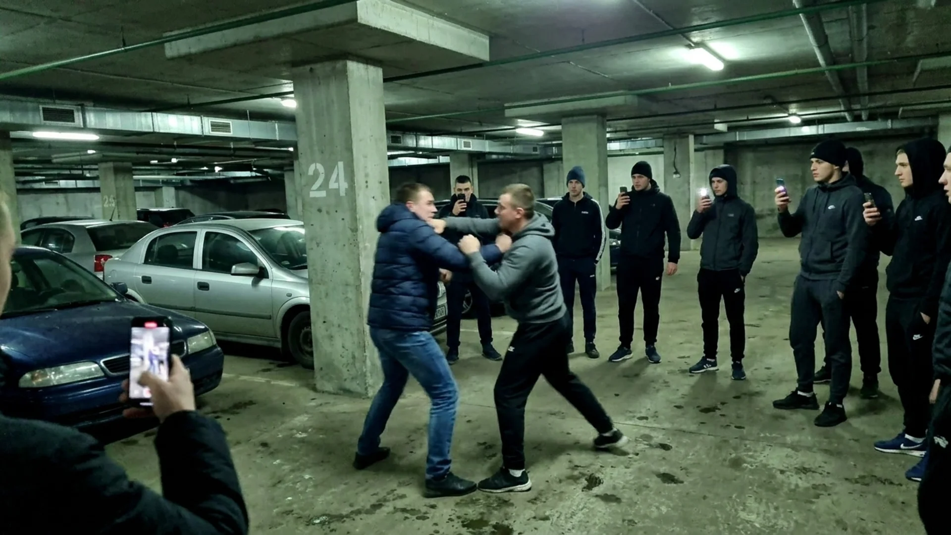 Two bare-knuckle fighters trade punches on the bare concrete floor of an underground car park while spectators film on phones